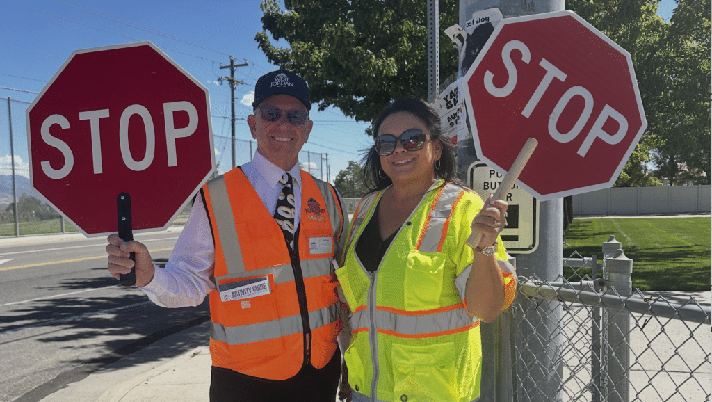 Two people wearing safety vests stand outdoors. One holds a small stop sign. They are both smiling, with shades and hats on under a sunny day. A fence, trees, and traffic signs are in the background.